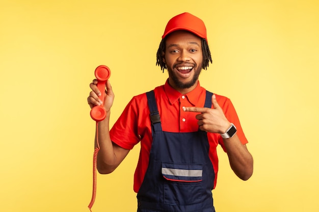 Trabajador barbudo satisfecho con uniforme sosteniendo un teléfono fijo rojo apuntando al teléfono esperando órdenes listas para trabajar Toma de estudio interior aislada en fondo amarillo