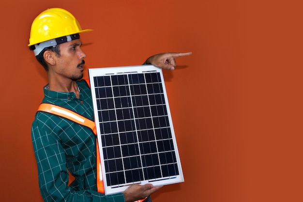 Trabajador con casco y equipo de protección individual sosteniendo un panel solar fotovoltaico aislado Trabajador solar aislado en fondo naranja