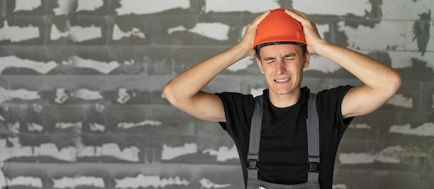 Trabajador con casco naranja cerca de un muro de piedras. Se tomó la cabeza con las manos. Copia espacio