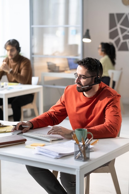 Trabajador del centro de llamadas con auriculares hablando con los clientes usando la computadora mientras está sentado en la mesa en la oficina