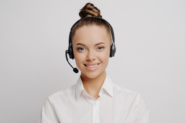 Trabajador del centro de llamadas sonriente con camisa blanca, cabello en pan, auriculares negros, ansioso por servir y ayudar a los clientes, de pie solo contra un fondo gris.