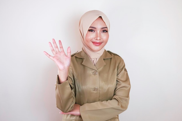 Trabajador civil musulmán con uniforme marrón y hijab saludando con un gesto de mano y sonriendo a la cámara