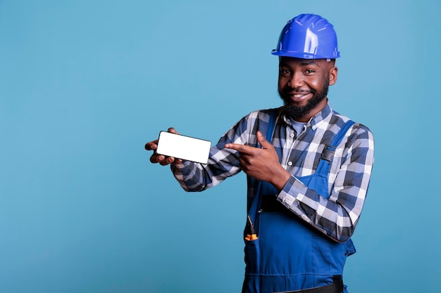 Trabajador de la construcción afroamericano con casco protector apuntando con el dedo al teléfono celular con pantalla vacía aislada en el fondo azul. Constructor en foto de estudio con pantalla en blanco de publicidad.