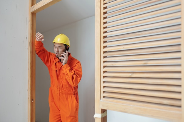 Foto trabajador de la construcción apoyado en el marco de la puerta y hablando por teléfono con un compañero de trabajo o un miembro de la familia