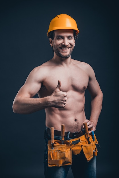 Trabajador de la construcción atractivo. Joven confiado gesticulando y mirando a la cámara mientras está de pie