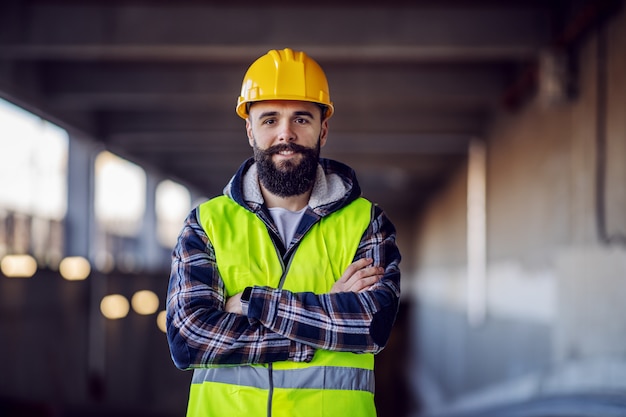 Trabajador de la construcción barbudo caucásico lindo con casco de seguridad en la cabeza en chaleco de pie con los brazos cruzados en el sitio de construcción