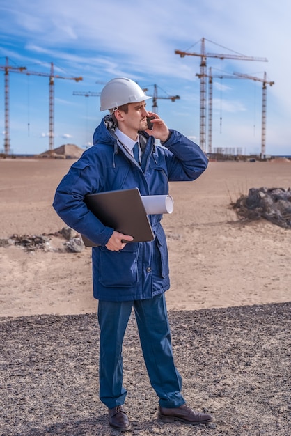 Un trabajador de la construcción en un casco blanco con una computadora portátil y documentos debajo del brazo