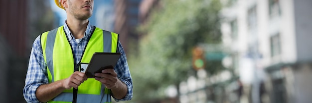 Trabajador de la construcción concentrado con tableta contra la vista borrosa de una ciudad moderna