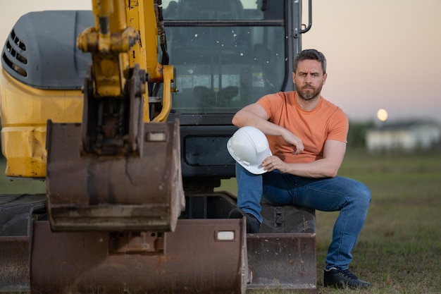 Trabajador de la construcción de hombre caucásico en casco en el sitio de construcción Trabajador de ingeniero industrial en casco cerca de excavadora o tractor excavadora Concepto de industria de la construcción Gerente de sitio de construcción