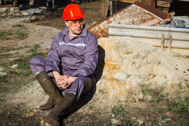 Un trabajador de la construcción joven en un casco naranja y gafas protectoras se sienta en una pila de arena.
