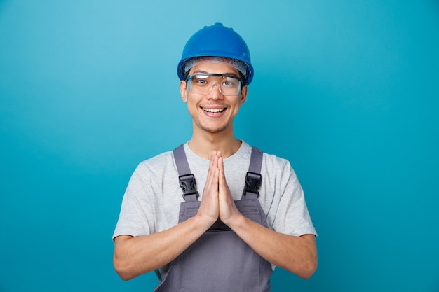 Trabajador de la construcción joven emocionado con casco de seguridad y uniforme con gafas de seguridad haciendo gesto de namaste