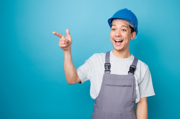 Trabajador de la construcción joven emocionado con casco de seguridad y uniforme mirando y apuntando a un lado