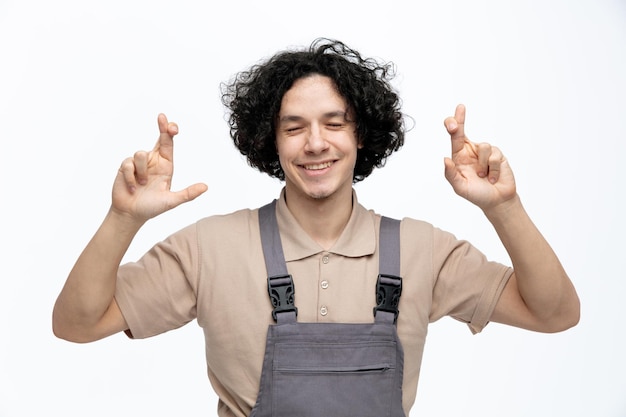 Trabajador de la construcción joven esperanzado vistiendo uniforme sonriendo mientras cruza los dedos con los ojos cerrados aislado sobre fondo blanco.