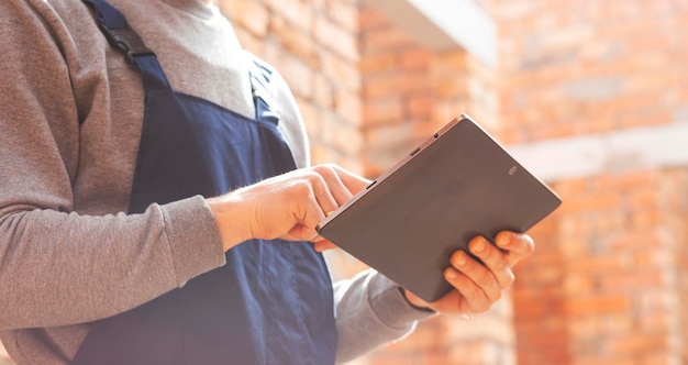 Trabajador de la construcción de pie en la casa en construcción y sosteniendo una tableta