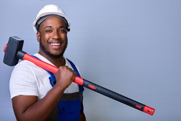 Trabajador de la construcción profesional con casco protector y ropa de trabajo mira a la cámara, sonríe y sostiene un mazo en el hombro Retrato de un trabajador negro con monos