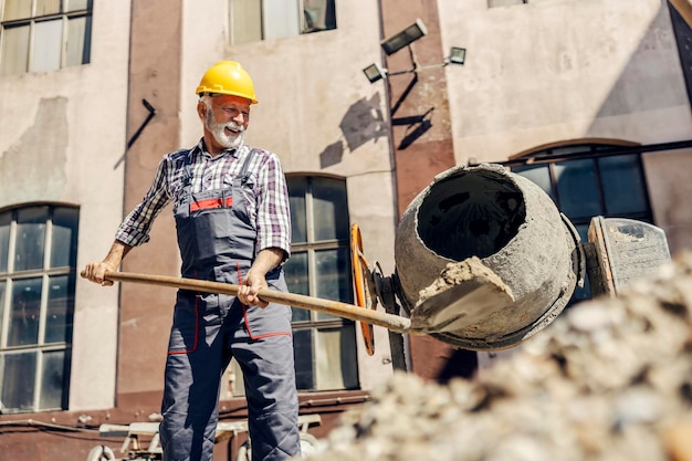 Un trabajador de la construcción senior está poniendo cemento en una licuadora de cemento mientras está de pie en el sitio