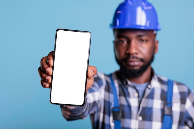 Trabajador de la construcción serio que muestra un teléfono moderno con pantalla blanca en blanco, maqueta publicitaria. Constructor afroamericano sosteniendo teléfono con pantalla vacía para promoción de aplicaciones.