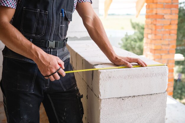 El trabajador de la construcción en el sitio de construcción mide la longitud de la abertura de la ventana y la pared de ladrillo con cinta métrica. La cabaña está hecha de bloques de hormigón poroso, ropa de trabajo, mono y gorra de béisbol.