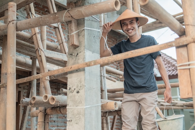 Trabajador de la construcción sonríe a la cámara mientras se para entre el bambú y los postes en la casa sin terminar