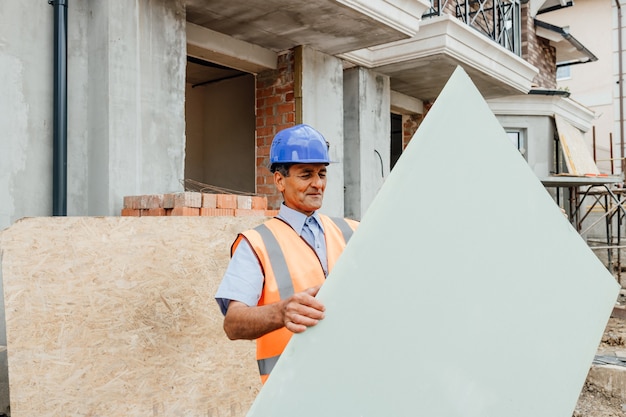 Un trabajador de la construcción sonriente en casco y chaleco sostiene un constructor de construcción de paneles de yeso de cartón sonriendo a la cámara copia espacio