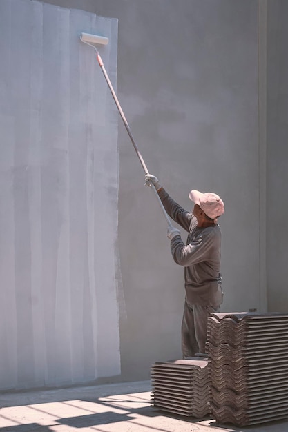 Trabajador constructor asiático aplicando pintura blanca de imprimación en el muro de hormigón dentro del sitio de construcción de la casa