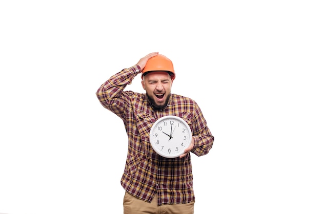 Trabajador constructor enojado y conmocionado en casco naranja de construcción protectora sosteniendo en la mano un gran reloj de alarma aislado sobre fondo blanco. Tiempo de trabajar. Tiempo de construcción de edificios.