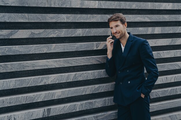 Trabajador corporativo masculino vestido con traje negro formal mantiene la mano en el bolsillo tiene conversación telefónica