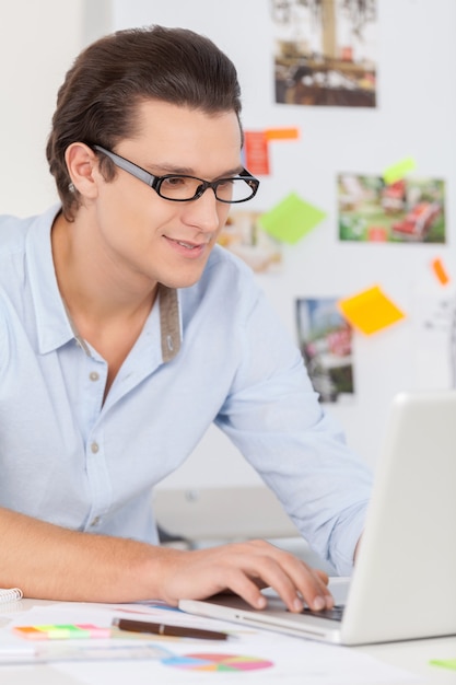 Trabajador creativo. Hombre joven alegre con gafas trabajando en la computadora y sonriendo mientras está sentado en su lugar de trabajo