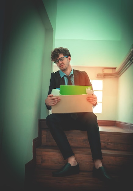 Foto trabajador desempleado que lleva una caja empacada sentado en la escalera de la oficina con expresión deprimida por concepto de desempleo