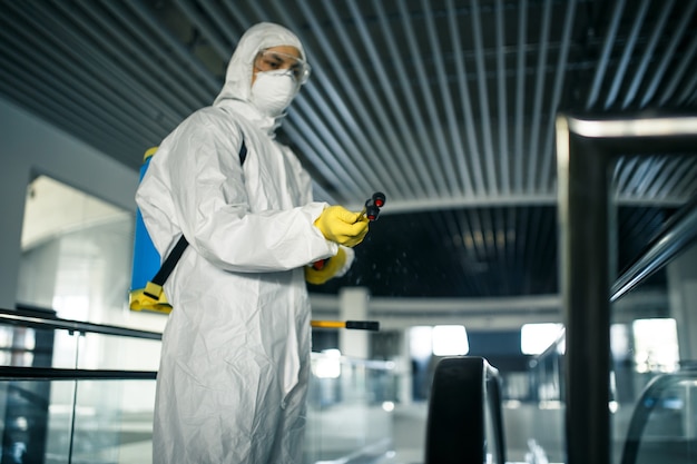 Trabajador de desinfección con traje protector limpia profesionalmente una escalera mecánica en un centro de negocios vacío. Un hombre equipado con ropa antibacteriana esteriliza el centro comercial. Concepto Covid.