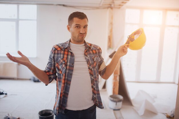 Trabajador divertido con casco de construcción Está de pie en el sitio de construcción y no sabe qué hacer.