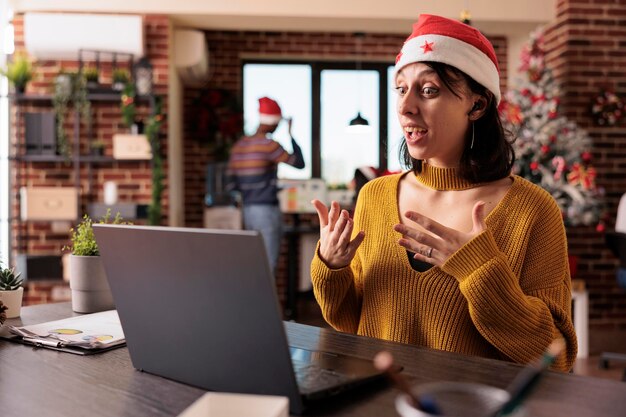 Trabajador de la empresa hablando en videoconferencia en la oficina con árbol de navidad y decoraciones festivas. Asistir a llamadas de teleconferencia remotas y reuniones en línea durante la temporada de vacaciones en el trabajo.