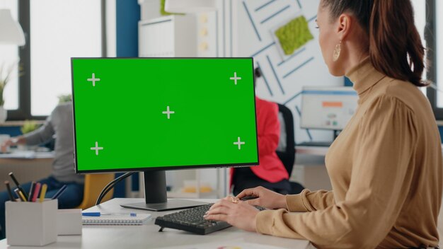 Trabajador de la empresa que usa una pantalla verde aislada en el monitor con clave de croma. Mujer de negocios mirando la computadora con plantilla simulada y antecedentes en la oficina de inicio. Espacio de copia de maqueta