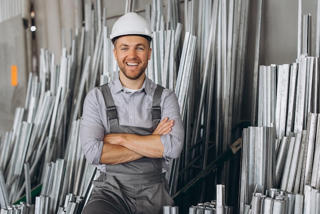 Foto trabajador de fábrica barbudo feliz con uniforme especial y casco blanco con marco de aluminio en la producción de ventanas y puertas de plástico metálico