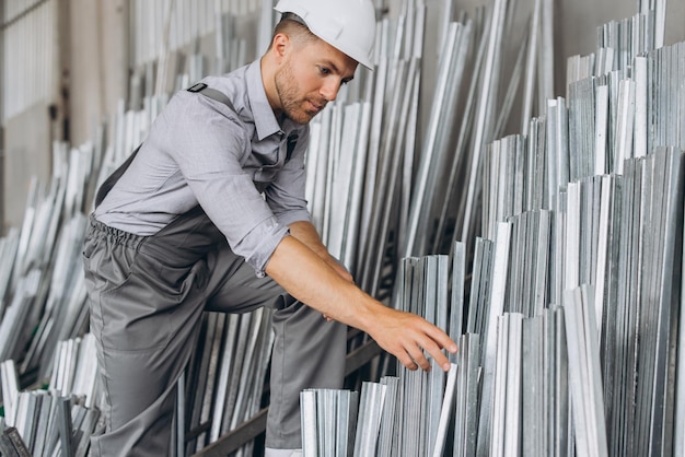 Trabajador de fábrica barbudo feliz con uniforme especial y casco blanco con marco de aluminio en la producción de ventanas y puertas de plástico metálico