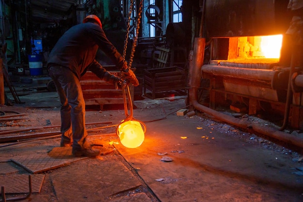 Trabajador de la fábrica de forja moviendo la pieza de trabajo de metal caliente brillante con focreps de grúa de cadena