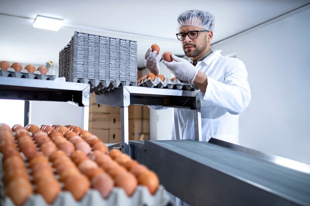 Trabajador de la fábrica sosteniendo huevos y de pie junto a la máquina de envasado en la granja de alimentos.