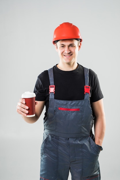 Trabajador feliz en casco con taza de papel con café para llevar y sonriendo a la cámara