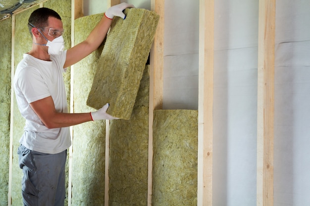 Trabajador en gafas protectoras y respirador aislante aislamiento de lana de roca en marco de madera para futuras paredes de la casa para barrera fría. Confortable casa cálida, economía, construcción y concepto de renovación.