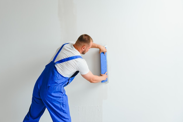 Un trabajador general en overoles usa una paleta para cubrir la pared con cemento.