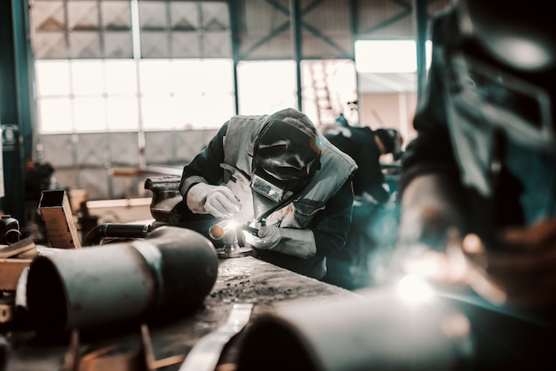 Trabajador de hierro en traje de protección, máscara y guantes de soldadura de tubería. Interior del taller.