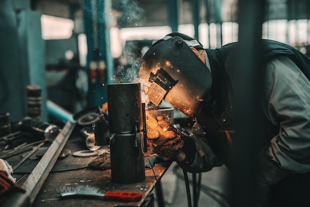 Foto trabajador de hierro en traje de protección, máscara y guantes de soldadura de tubería. interior del taller.