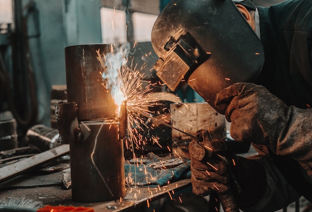 Trabajador de hierro en traje de protección, máscara y guantes de soldadura de tubería. Interior del taller.