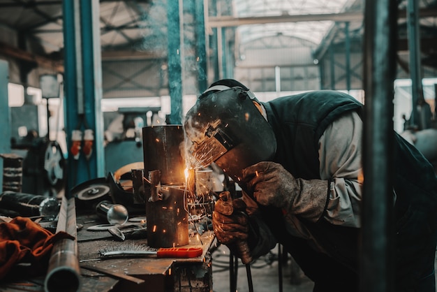 Trabajador de hierro en traje de protección, máscara y guantes de soldadura de tubería. Interior del taller.