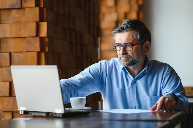 El trabajador independiente masculino está trabajando en un café en un nuevo proyecto empresarial Se sienta en una ventana grande en la mesa Mira la pantalla de una computadora portátil con una taza de café