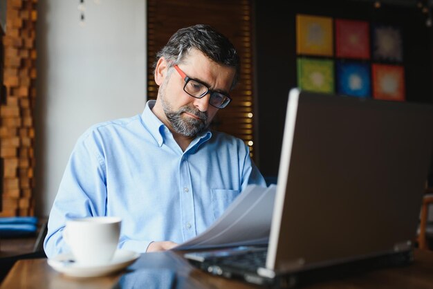 El trabajador independiente masculino está trabajando en un café en un nuevo proyecto empresarial Se sienta en una ventana grande en la mesa Mira la pantalla de una computadora portátil con una taza de café