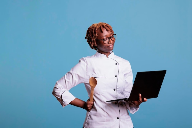 Trabajador de la industria alimentaria con uniforme de cocina usando una computadora para inspirar una comida mediterránea. Chef con laptop buscando recetas de platos en internet mientras está de pie en el estudio.