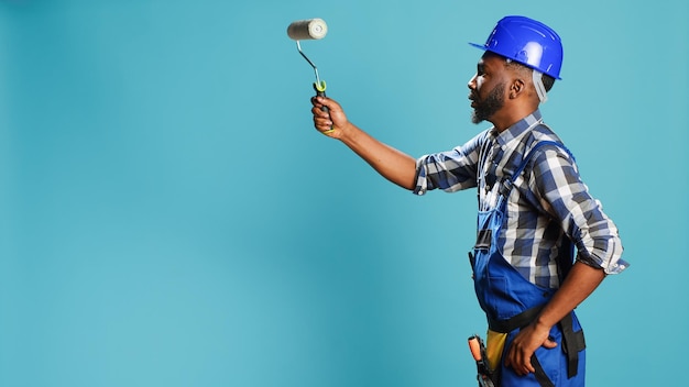 Trabajador industrial en uniforme pintando paredes con rodillo, usando un instrumento de renovación sobre fondo azul. Artesano profesional con casco trabajando con pincel rodante para cambiar de color.