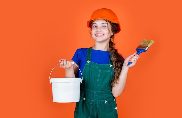 Trabajador infantil alegre con uniforme de construcción y cubo de pintura y pincel, trabajo creativo.