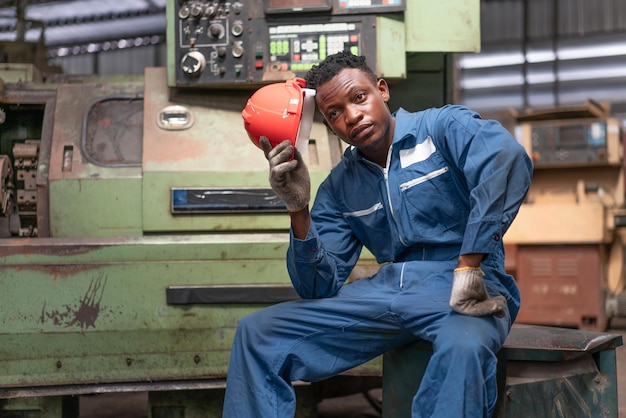 Trabajador ingeniero afroamericano sentado en una máquina vieja sintiéndose cansado del trabajo duro en la fábrica